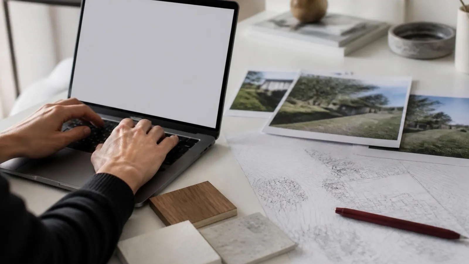 Architecte qui rédige une notice sur un ordinateur portable, photos du terrain et plan de masse imprimés à côté.